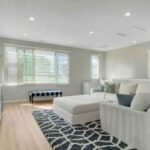 Bright living room with white sectional sofa, matching ottoman, a blue geometric rug, and large windows with blinds.